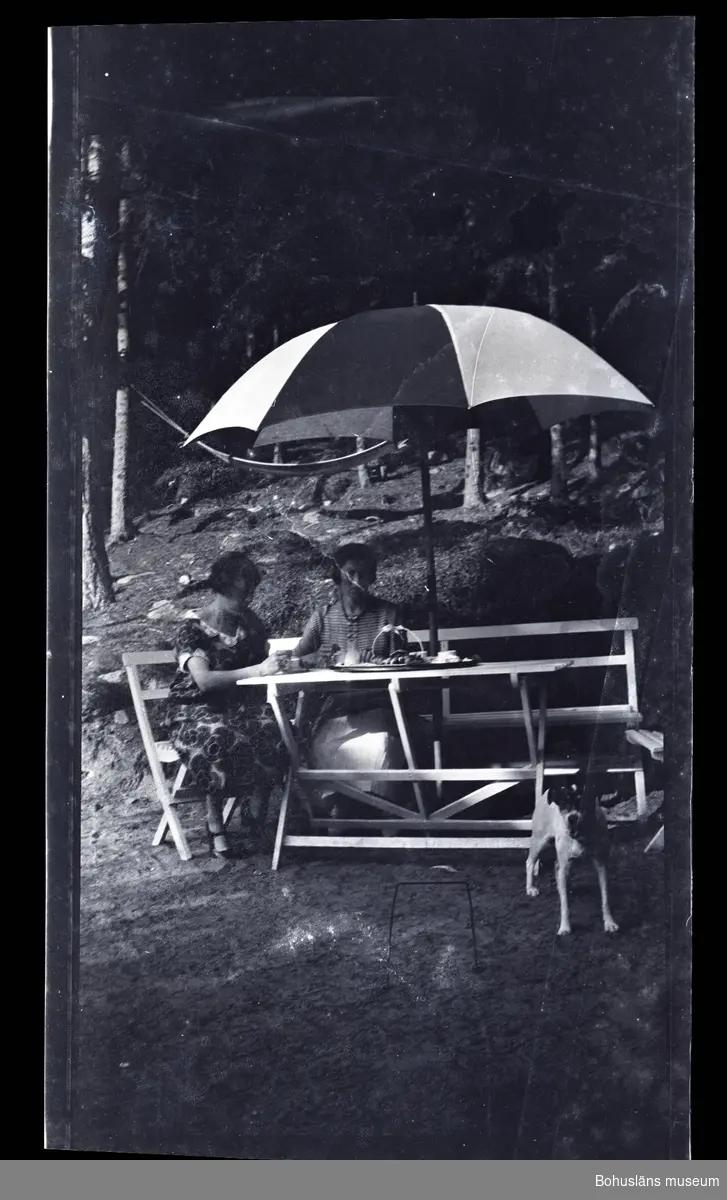 Två personer sittandes i trädgårdsmöbler under ett parasoll med en hund i förgrunden.