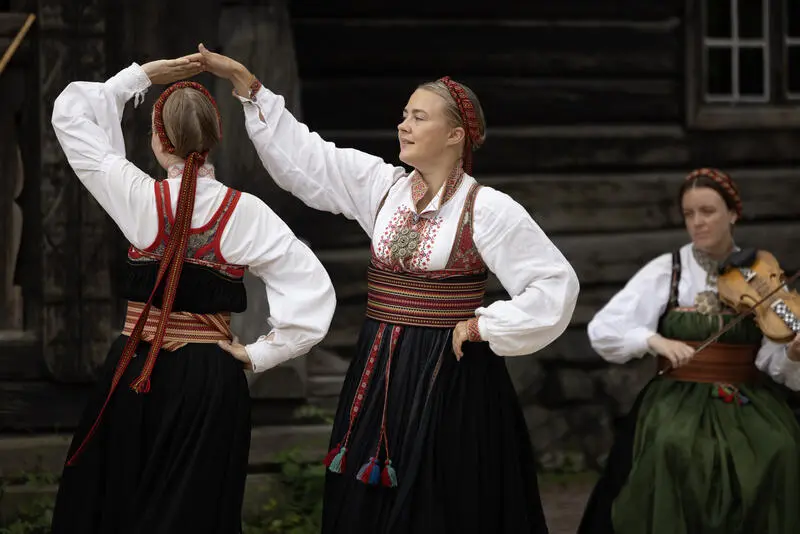 To kvinner i bunad danser folkedans, en tredje spiller fele.