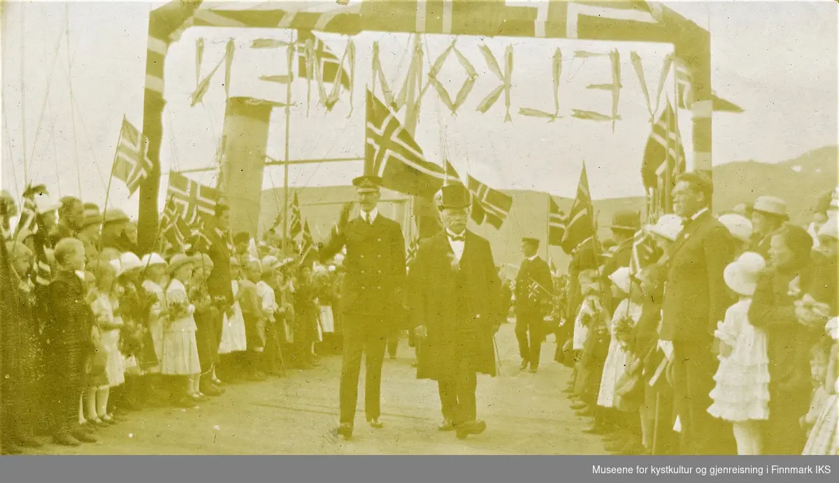 Kong Haakon VII ankommer Honningsvåg. Han passerer sammen med John Bruun velkomstportalen og ble møtt med flagg og hurra-rop. 28.06.1922.