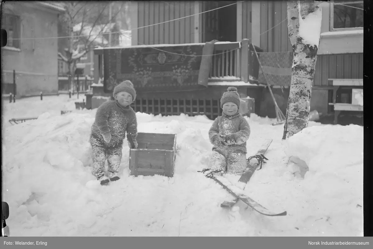 To barn kledd i strikkeplagg leker i snøen utenfor bolighus. Ett av barna holder seg i en trekasse, det andre sitter på kne foran ski med snørebindinger. Gulvteppe henger til lufting på veranda ved inngangsparti til bolighus. To bolighus, hvorav ett murhus i bakgrunnen.