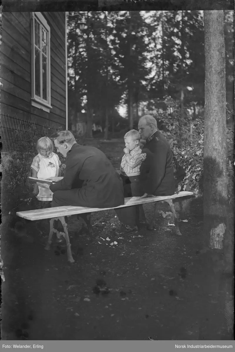 To menn sittende på benk kledd i Frelsesarmeenuniform. To barn stående foran. En av mennene viser en bok til jenta.