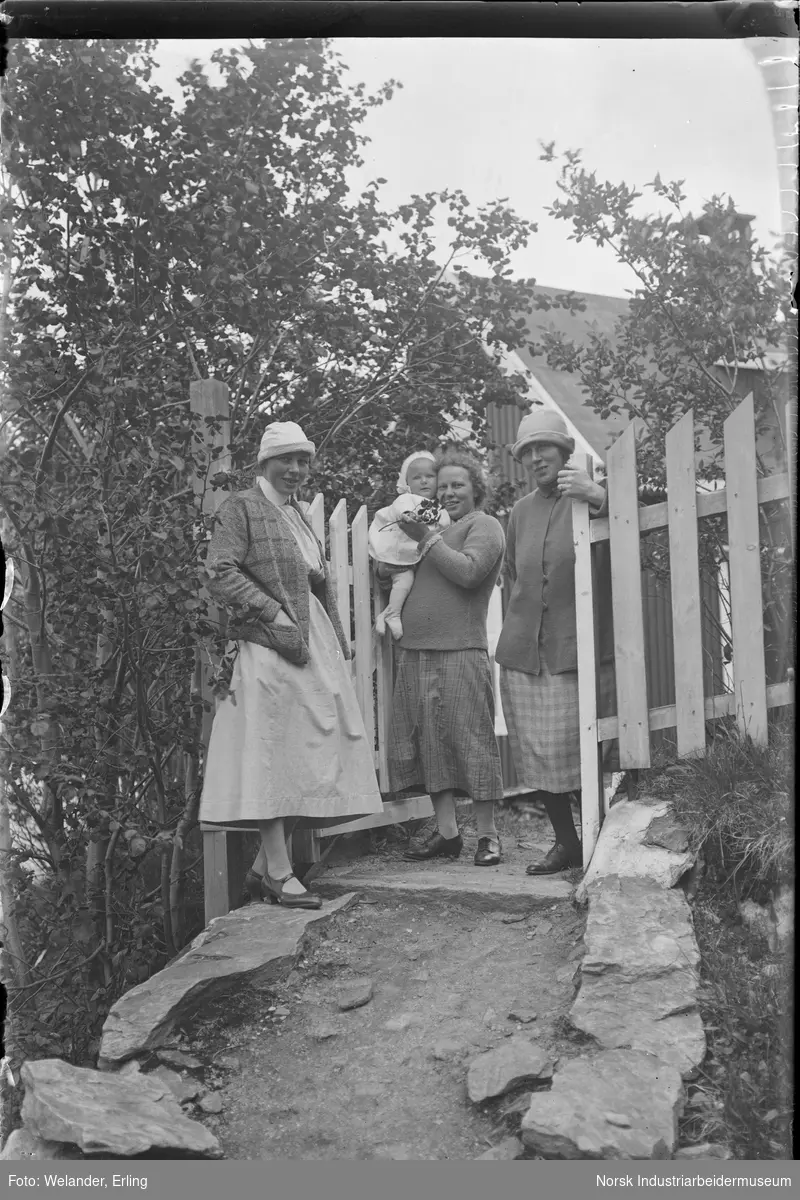 Tre kvinner, en av de med ett barn på armen stående ved inngangsporten til ett bolighus. Barnet har en blomst i hånden. To av kvinnene har bøttehatt på hodet.