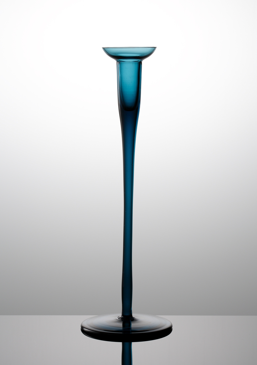 Formgivare: Nils Landberg. Stålblå hög ljusstake på fot. Cylindrisk ljushållare med halvsfärisk ljusmanschett. Draget ben med rund skivfot.