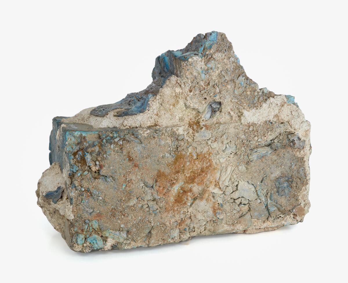 Byggsten av gjuten masugnsslagg. Troligen från järnbergslagen i södra Dalarna. Oregelbunden rektangulär form med utstickande delar som vittnar om att blocket en gång ingått i ett murverk. De höga temperaturerna vid järnframställningen har fått slaggens yttre skickt att omvandlas till glas. Kulörerna i detta skickt varierar kraftigt och beror på de mineraler som förekommer i malmen. Detta block är främst ljust- till mörkblått, vilket tyder på halter av kobolt och krom.