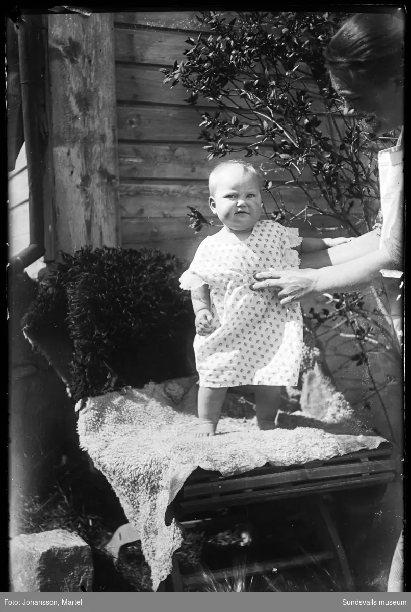 Ett litet barn i ettårsåldern står på en fäll utomhus. En kvinna hjälper den lilla att hålla balansen.