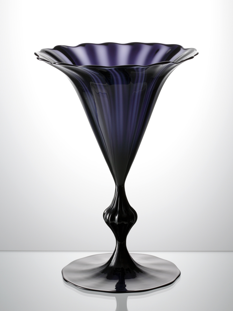 "Slottsglas" formgivet av Simon Gate till Göteborgsutställningen 1923. Friblåst och räfflat i lila glas. Fot och kupa blåsta och drivna. Räfflat ben med en väl tilltagen knapp.