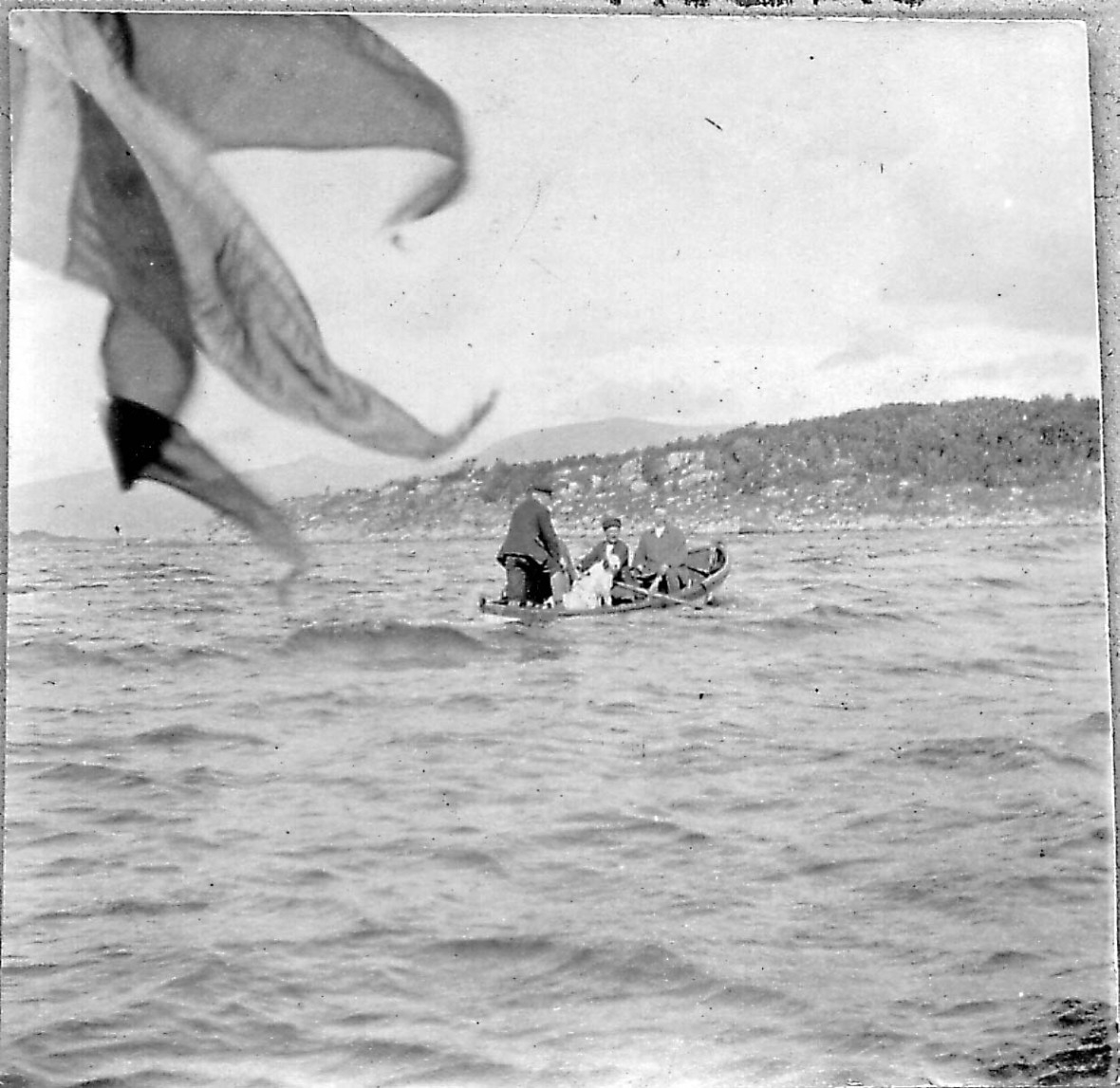 Tre menn i spissbåt med hund. Bildet er tatt fra en annen båt