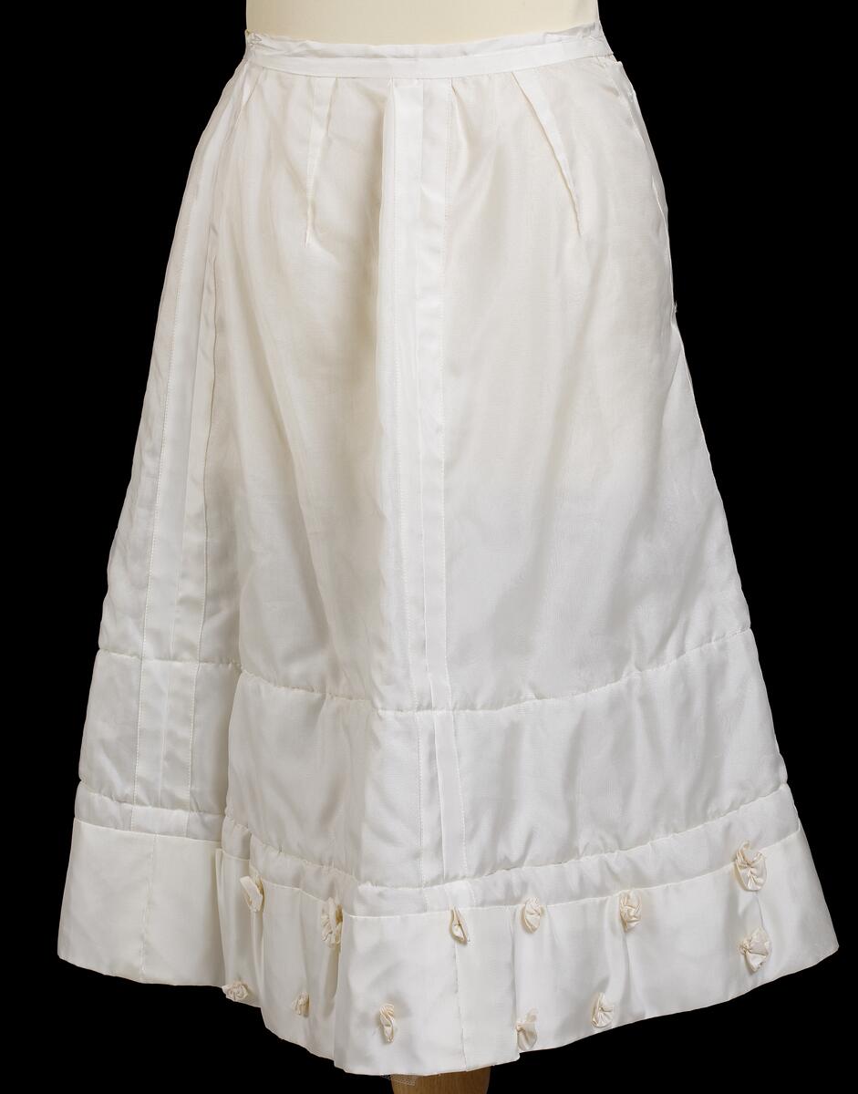 Underkjol till brudklänning i vitt konstfibermaterial. Längst ner på kjolens insida en rand av tyll. Hela brudklänningen består av klänning, underkjol, jacka, skor, sidenband, handskar och brudkrona.