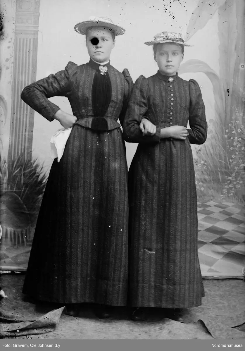 Portrett i helfigur av to kvinner i kjole med hatt. Den ene kvinnen holder i et lommetørkle.