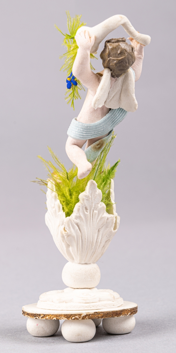 Bröllopskaramell, föreställande ängelfigur/gosse, som står med ymnighetshorn i öppna växtblad.