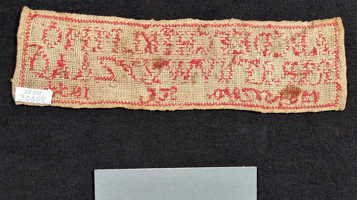 Märkduk med broderi i rött garn, alfabetet och siffror från 1-0. Tyget är av tuskaftat linne.