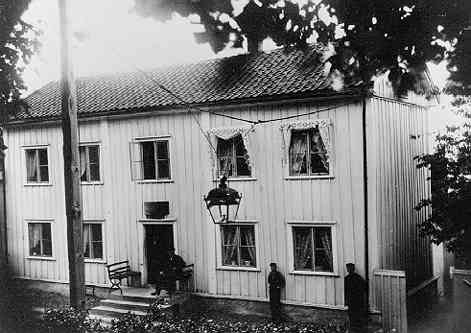 En man sitter i en soffa på farstun till en fastighet utmed Brahegatan i Gränna - en kvinna och en flicka står innanför dörren. Till höger om farstun står en pojke och en man lutad mot husväggen. På väggen ovanför farstudörren sitter en skylt med otydlig text, avser A F Hellströms gördelmakarverkstad e dyl.
En gatlykta hänger över gatan, tillverkad av 1848 av kopparslagare Holmgren.