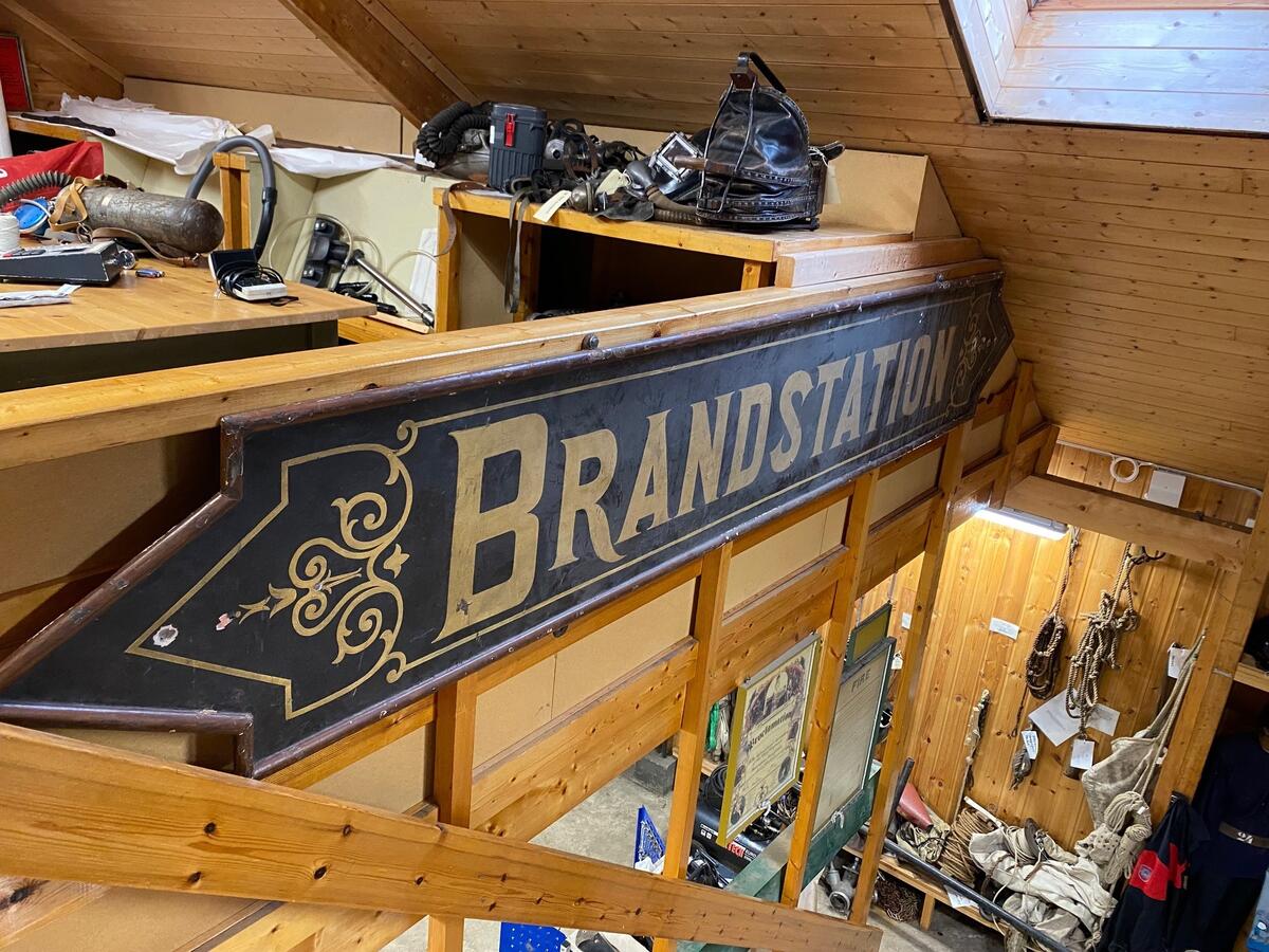 Skilt som har hengt på hovedfasaden på Gamle Hovedbrannstasjon i Bergen. Rektangulært med spisse ender på hver side. Påmalt "BRANDSTATION" med gullskrift på svart bakgrunn, brun kant rundt hele skiltet. Dekorert med gullornamentikk rundt, foran og etter skriften.