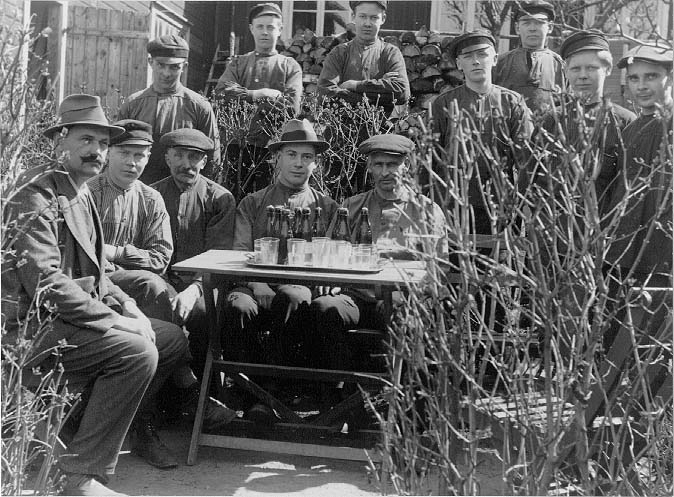 Tolv män och pojkar i arbetarkläder i en trädgård runt ett bord. På bordet står det läskedrycker och glas. Det är mannen till vänster, fabrikör A E Bolling som bjuder sina anställda vid hans ljuskronefabrik.