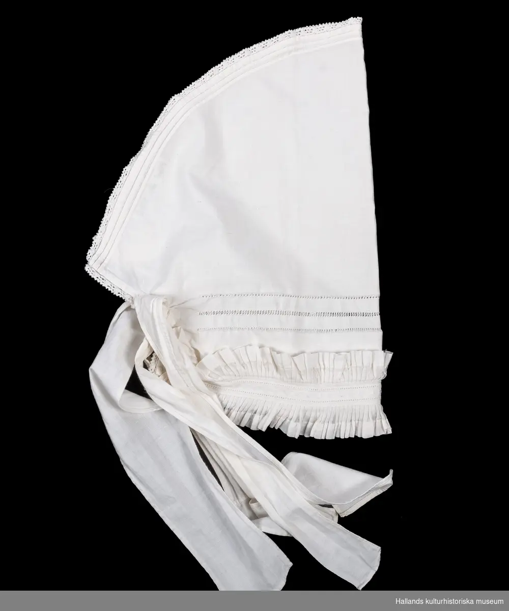 Huvudkläde av vit bomull med veckat krus framtill, hålsöm och spetskantat runt om. Två par knytband.