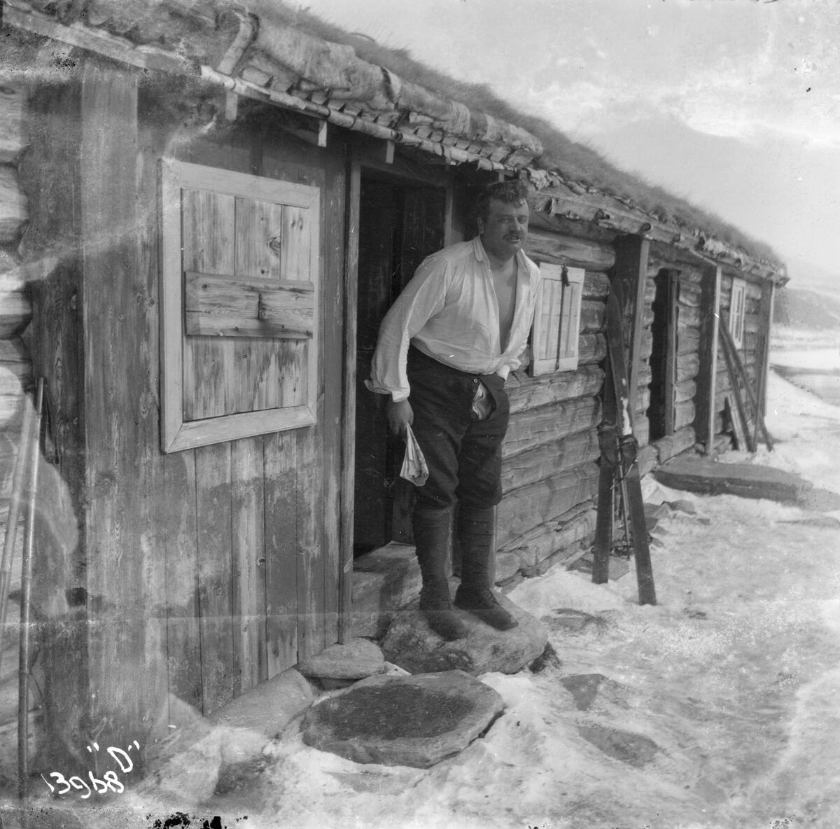 En mann står i døråpningen til en laftet tømmerhytte eller seterhus.