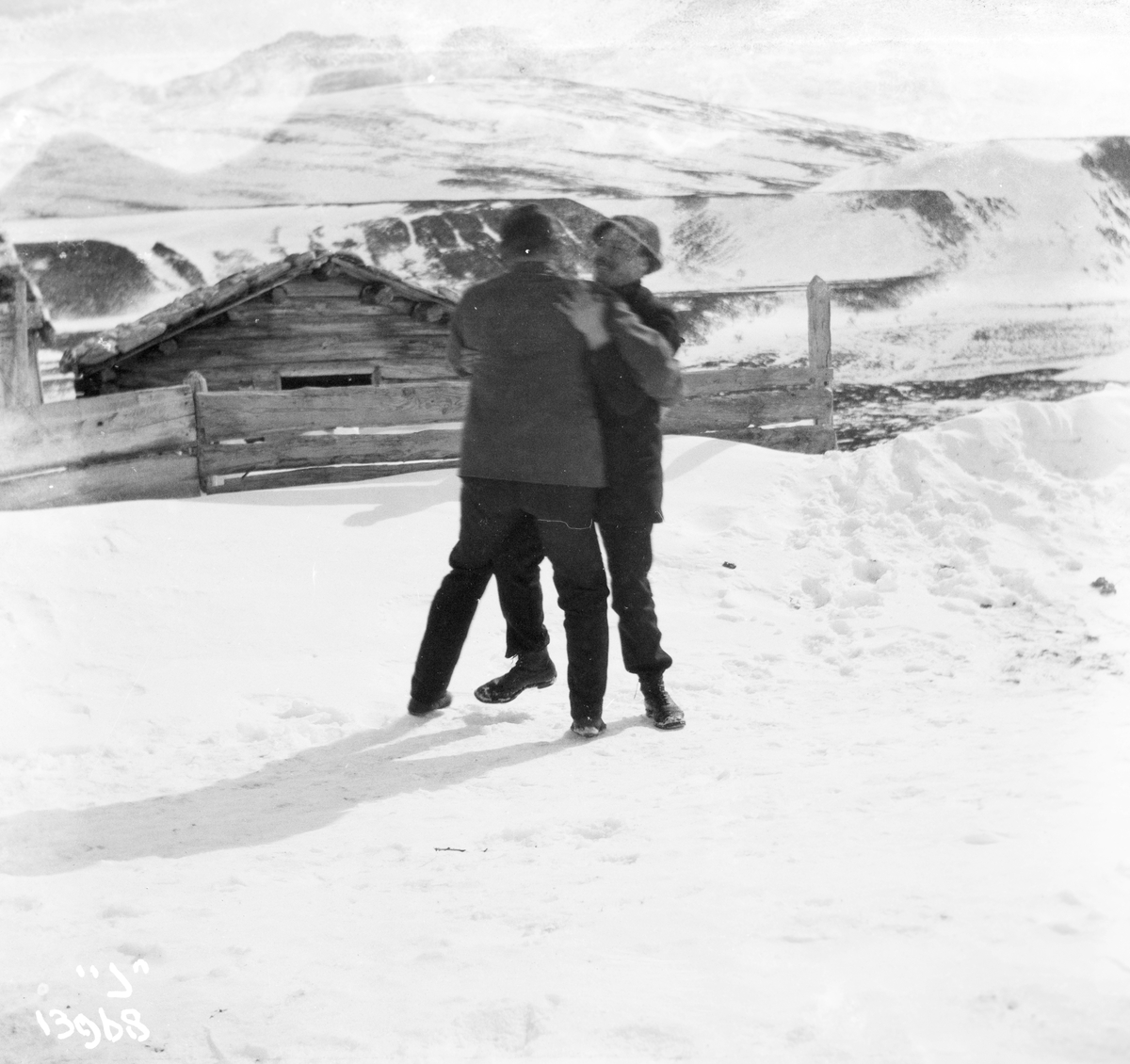 To menn danser i snøen utenfor en laftet tømmerhytte.