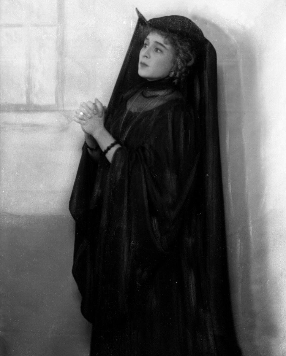 Portrett av skuespilleren og sceneinstruktøren Gerda Ring. Hun poserer i en svart kjole og en vid hodebekledning. Hun står oppreist og folder hendene.