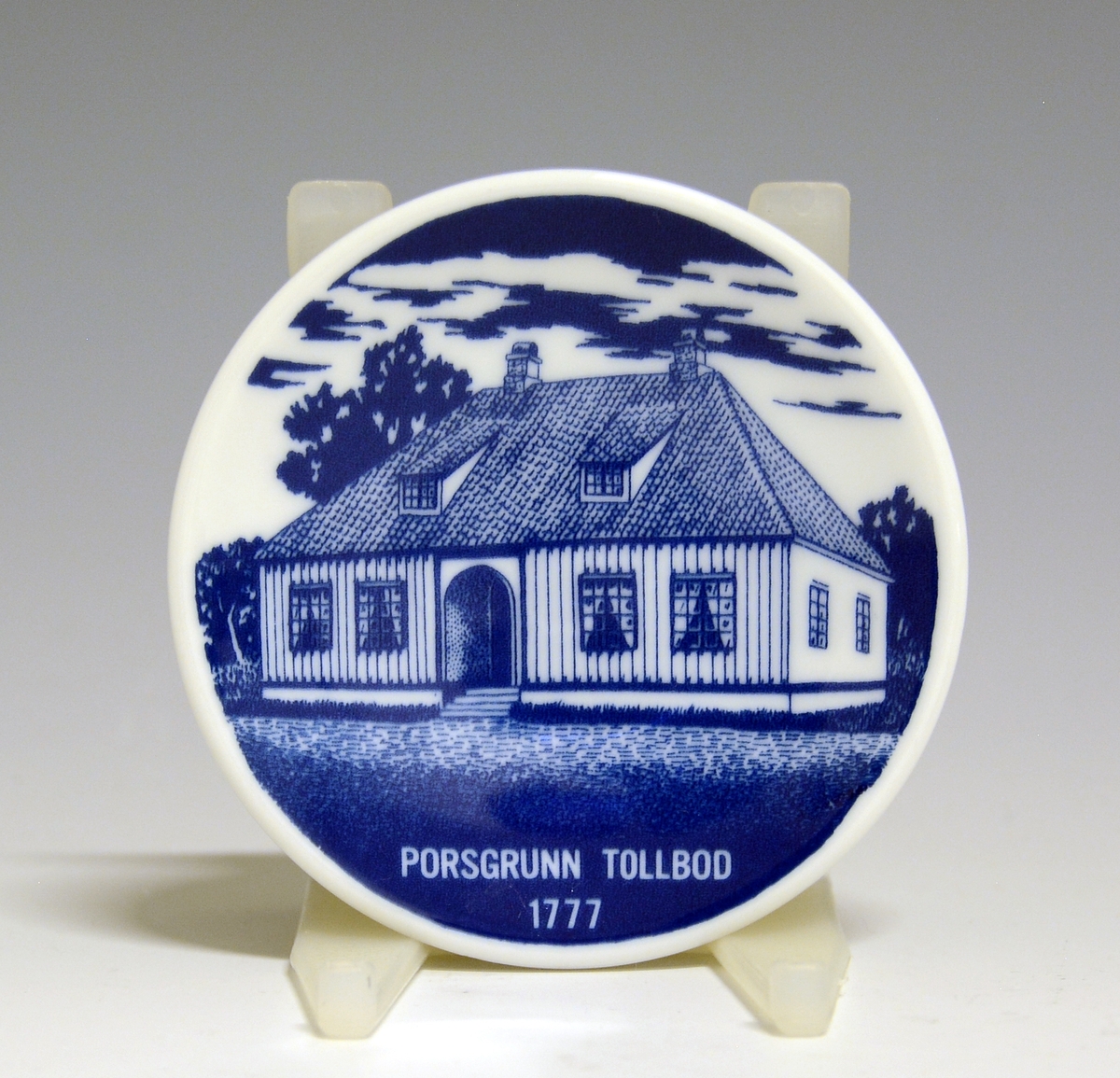 Miniatyrplatte av porselen. Hvit glasur. Motiv i blått av Porsgrunn Tollbod 1777.
Motiv av Harald Vemren.