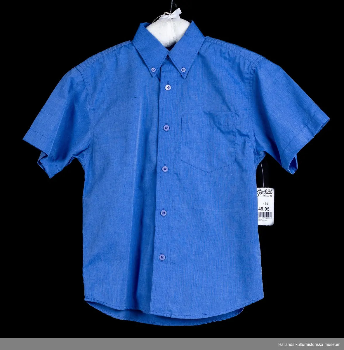 Kortärmad, klarblå skjorta med krage och en bröstficka. Knäpps med 6 blå knappar och har två blå kragknappar. Märket i nacken är "TIGERS CREW CLASSIC", Storlek 130 och priset är 49.95 kr. enligt prislappen. Längd ärm:180 mm.  