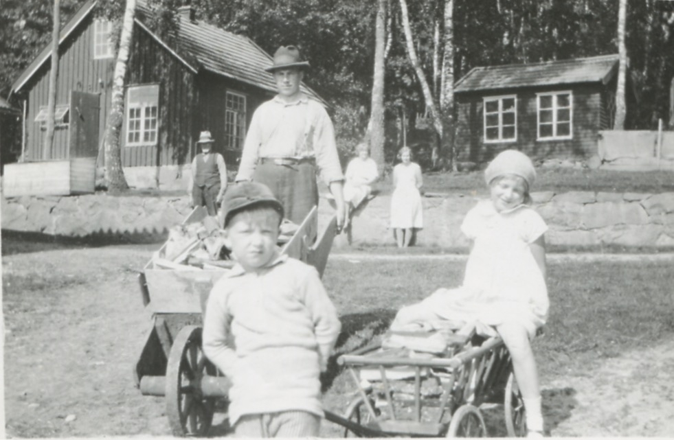 Vuxna och barn förflyttar ved i kärror, Kållered Stom "Nygård", 1930-tal. Ekonomibyggnader ses i bakgrunden.