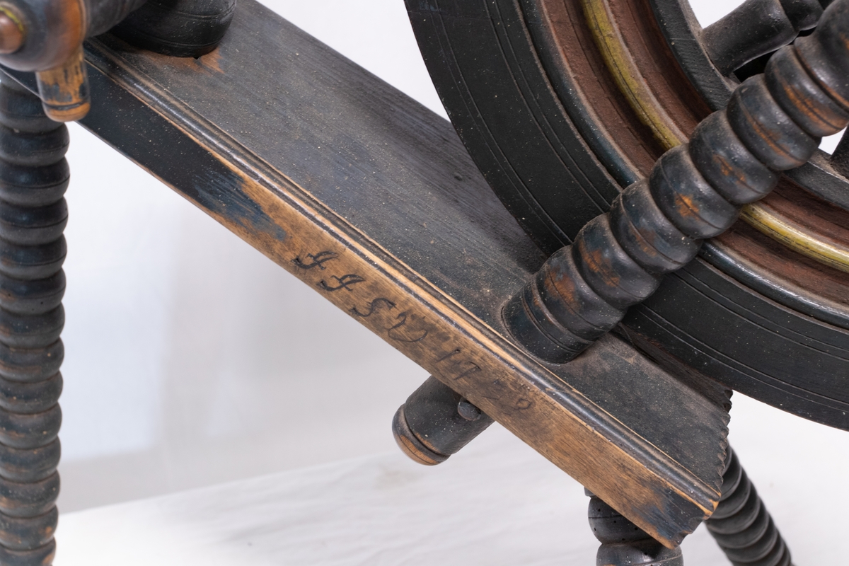 Grønnsort rokk med Trebeint skråttstillet krakk, der den ene ende er forsynt med skrue. Hjulet har tolv eker og bred rigg som henger med to skråstøtter. Forbindelse mellom hjul og pedal mangler.