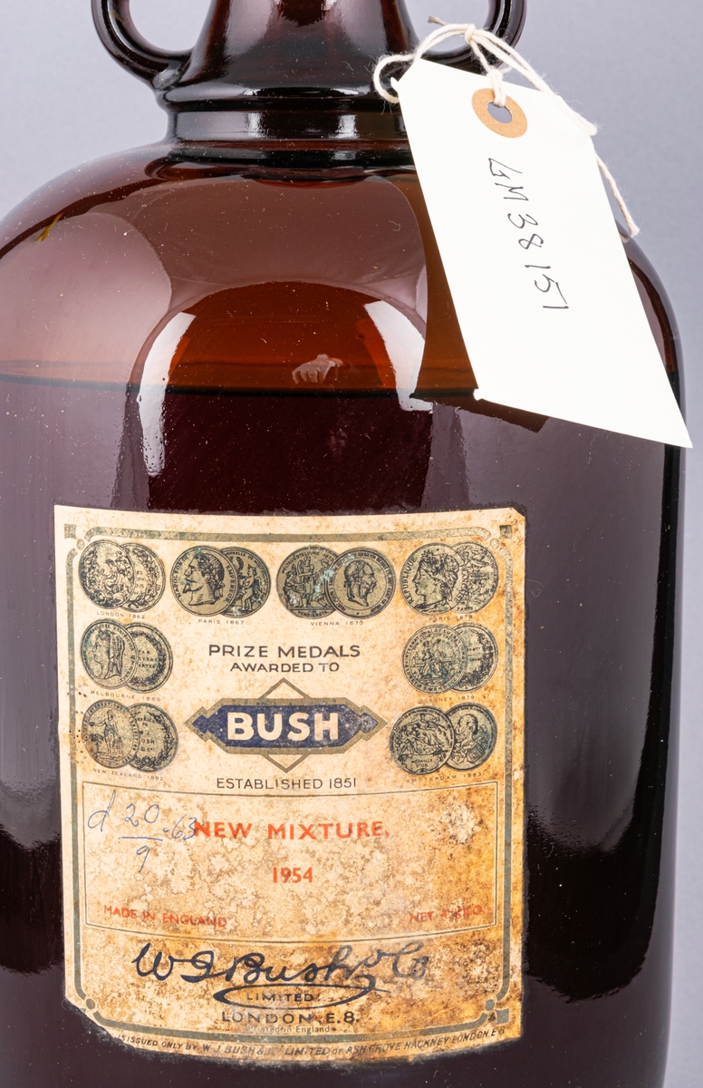 Glasflaska, brun, med hänklar och röd kork. Innehåller essens. Etikett med text: "BUSH new mixture 1954".