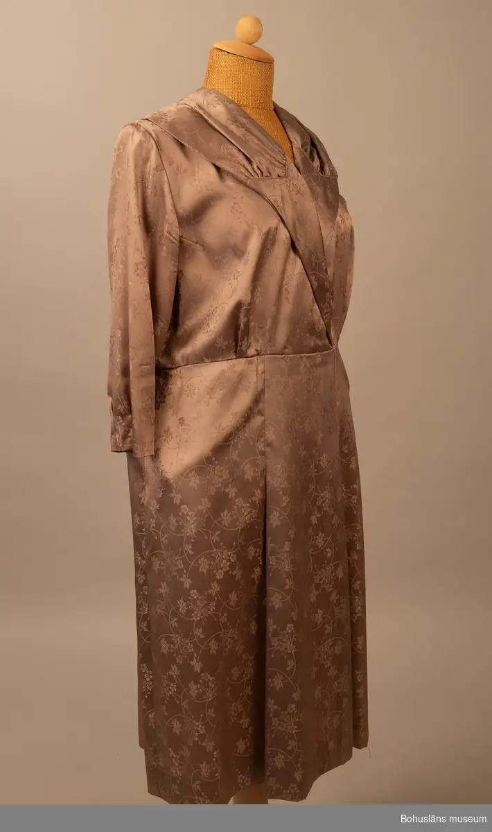 Klänning med schalkrage och halvlång ärm, avskuret liv och kjol med veck i grå syntetdamast. Dragkedja i sidan.
Användes på 1980-talet då det var mode med 1950-talsklänningar.

Ärmlängd 30cm, kjollängd 69cm.