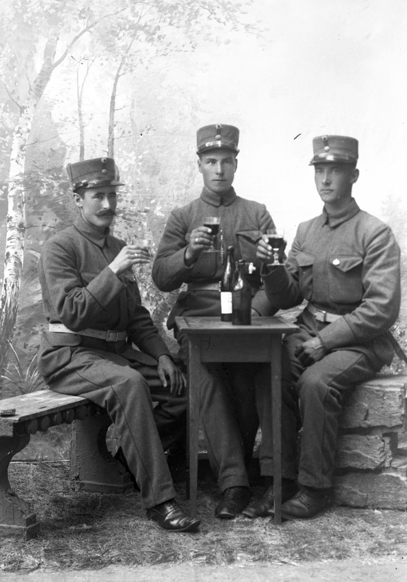 Atelierfoto.Tre soldater sitter ved bord med vinflasker på og glass med vin i hender.
