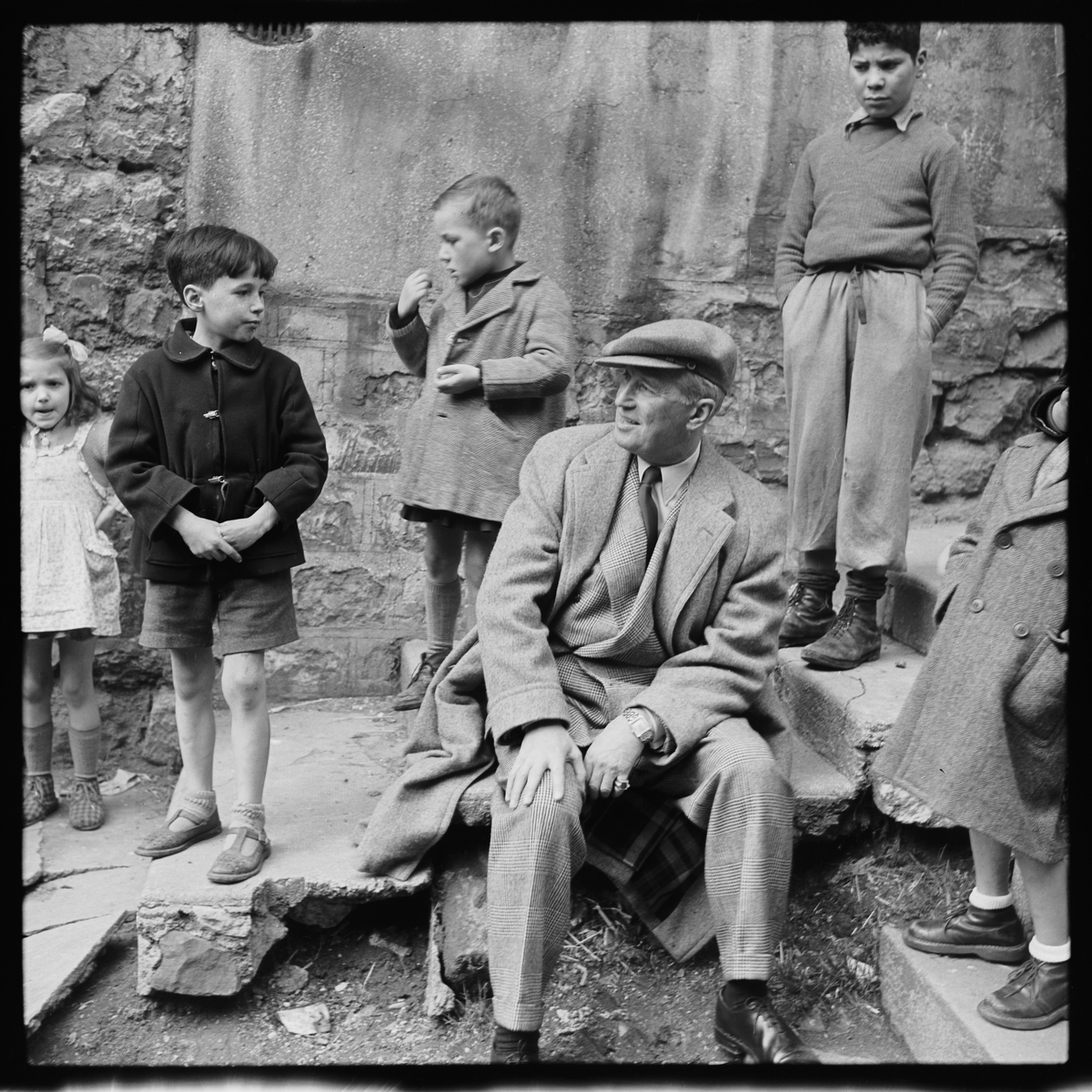 Maurice Chevalier sitter på en trapp, rundt han står tre gutter. 