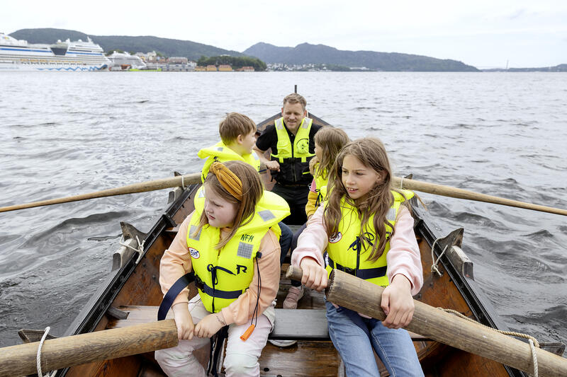 4 barn i en robåt under oppsyn av voksen, alle iført gule redningsvester.
