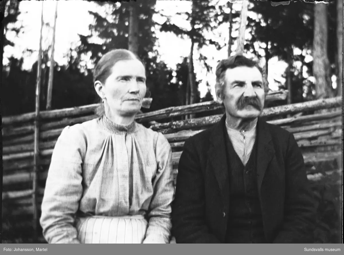Porträtt av makarna  Andrietta och Johan Persson, fotografens föräldrar, utomhus med en gärdesgård i bakgrunden.