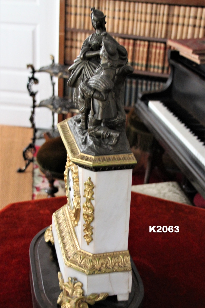a) Kaminur, fransk eller engelsk 1830-årene. Alabast og forgylt bronse. 
Ustemplet verk. Hvit emaljert tallskive med sorte romertall, omgitt av forgylt ramme. Avskåret ved hjørnene med bronseornamenter. På toppen knelende mann i bronse med sigd, kornnek og kvinne som gir ham drikke. 
Tilhørende to nøkler. 
b) Oval sokkel av Jakaranda med innlagt messingstripe.