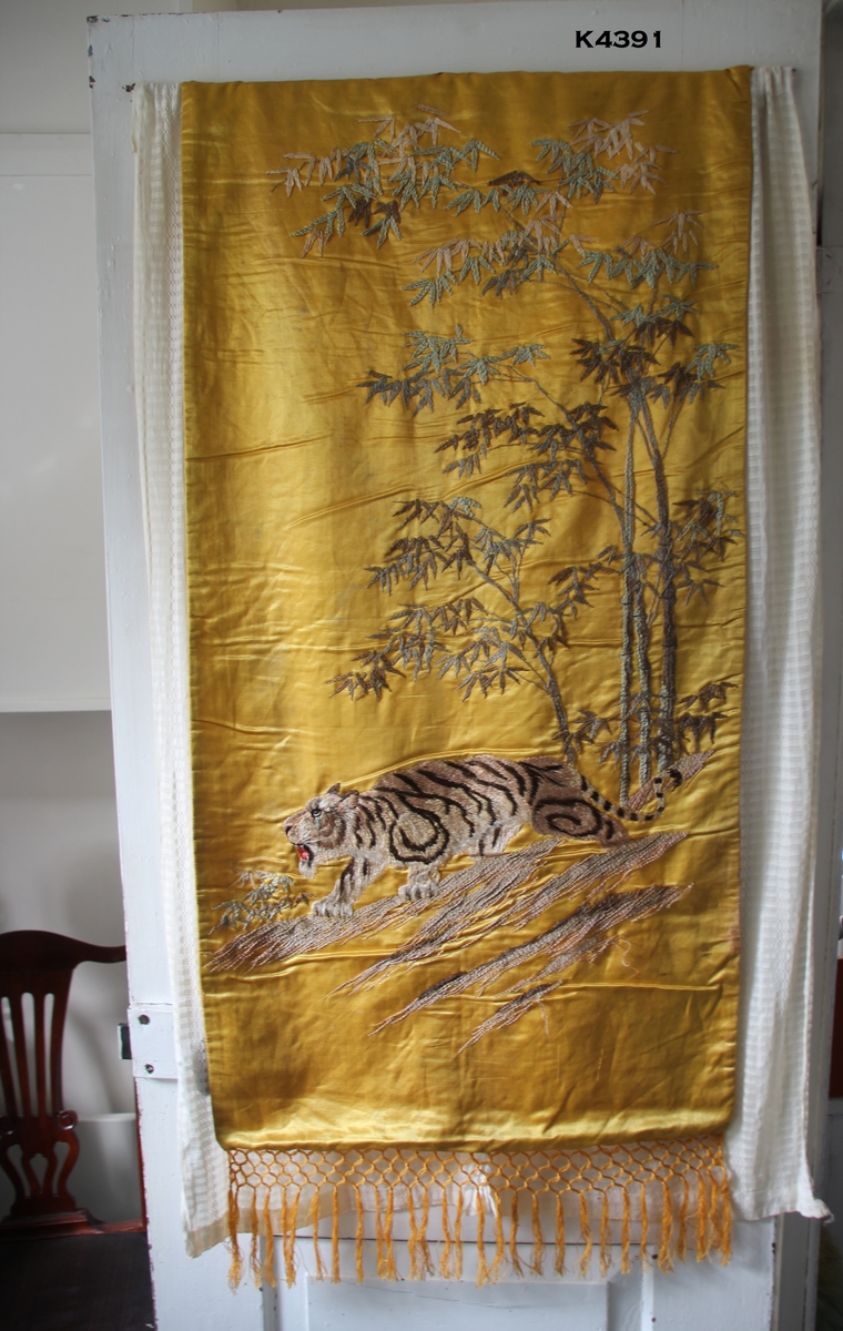 Silkebilde med motiv av en tiger, trær og kvister. Plattsøm, brodert med silketråd. Trær i grått og brunt. Tigeren har rød tunge og 4 tenner. Foret med gul silke. Frynser som er laget i silkegarn.
