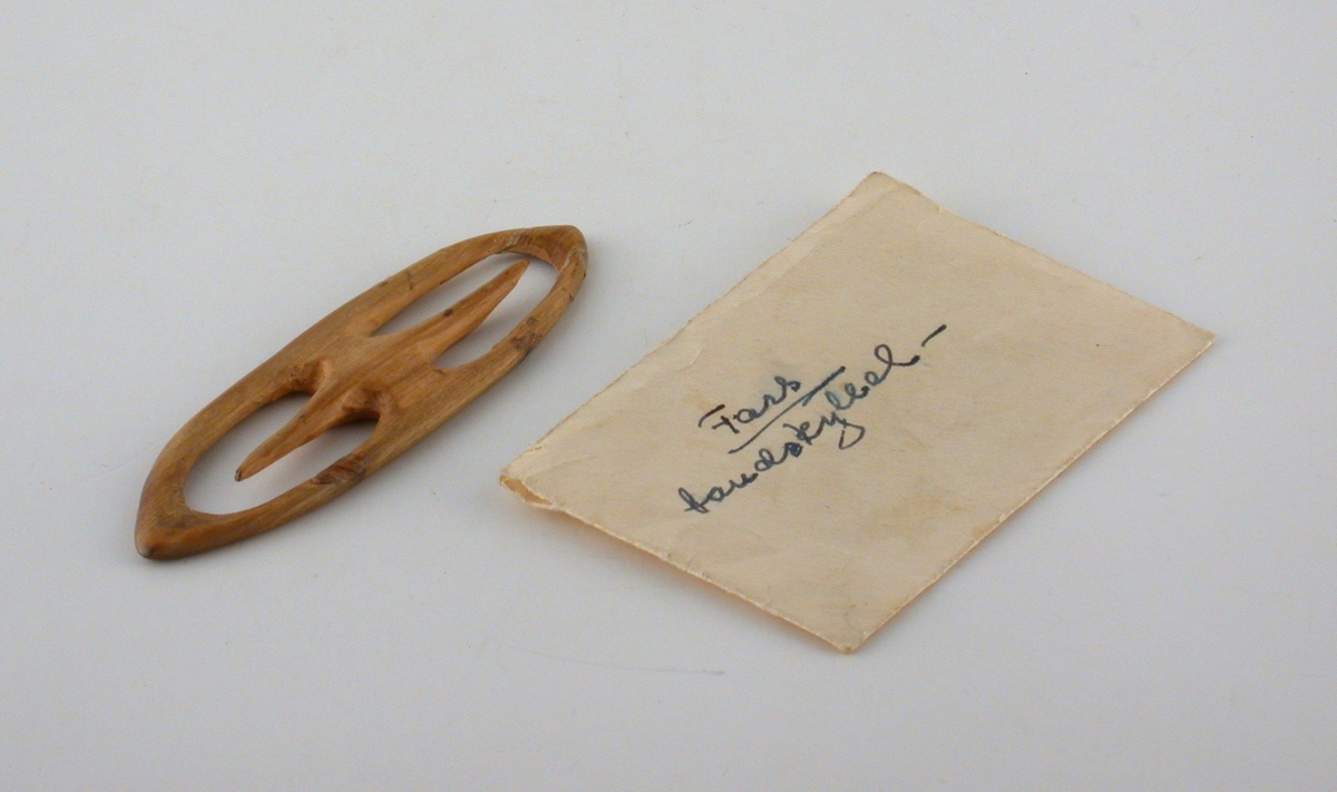 Handgjord bandskyttel i trä, oval form med utskurna lite vässade piggar från mitten (för att linda garnet runt). Tillhörande kuvert med texten "Fars bandskyttel".