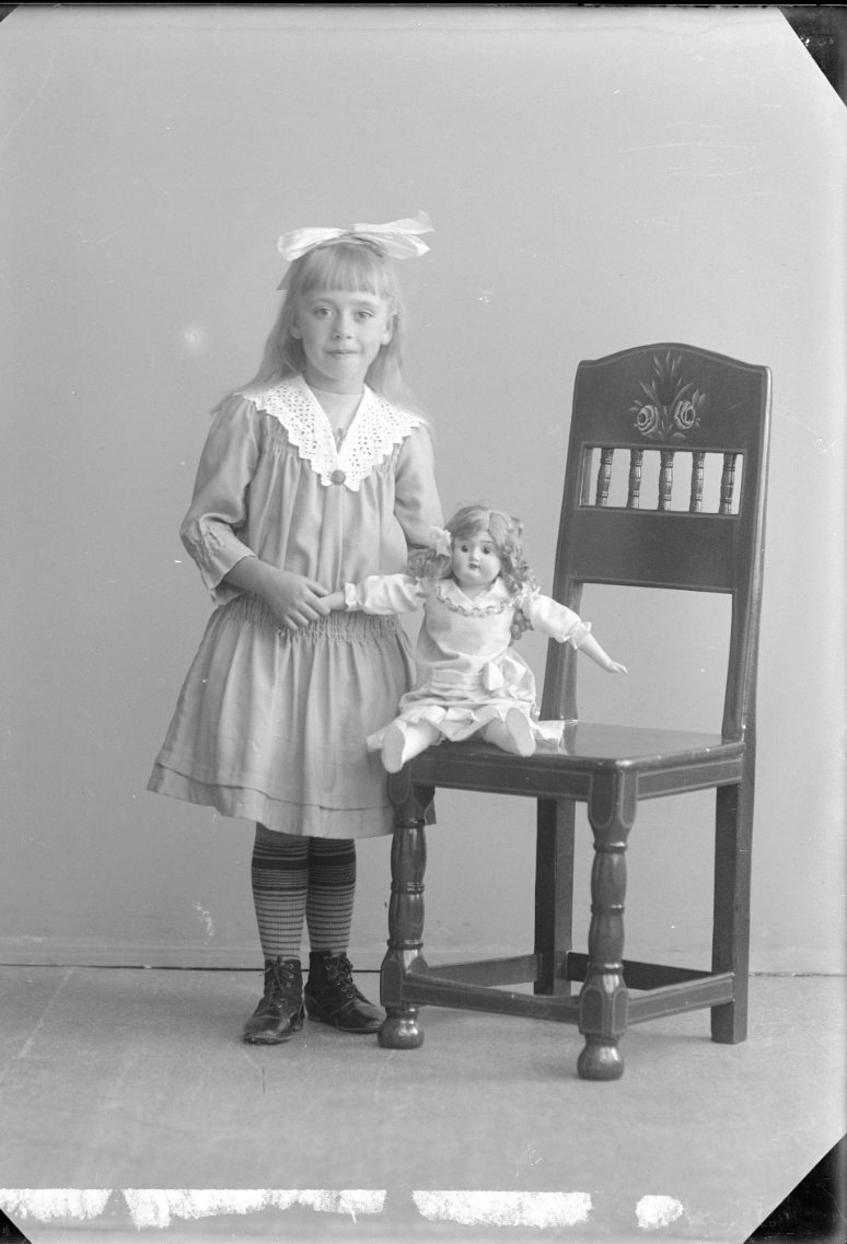 Porträtt av en flicka med rosett i håret. Hon står vid en stol med en docka som sitter på stolen.
