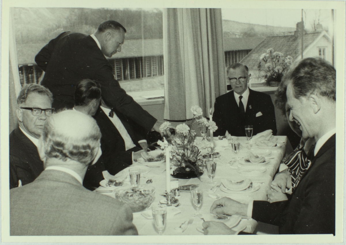 Finkledde kvinner og menn sit ved pynta langbord. Ved enden av bordet sit Sigurd J. Øglænd. Fotoet er frå Øglænd sitt 100-årsjubileum i 1968, truleg frå eit arrangement for kundar og leverandørar.