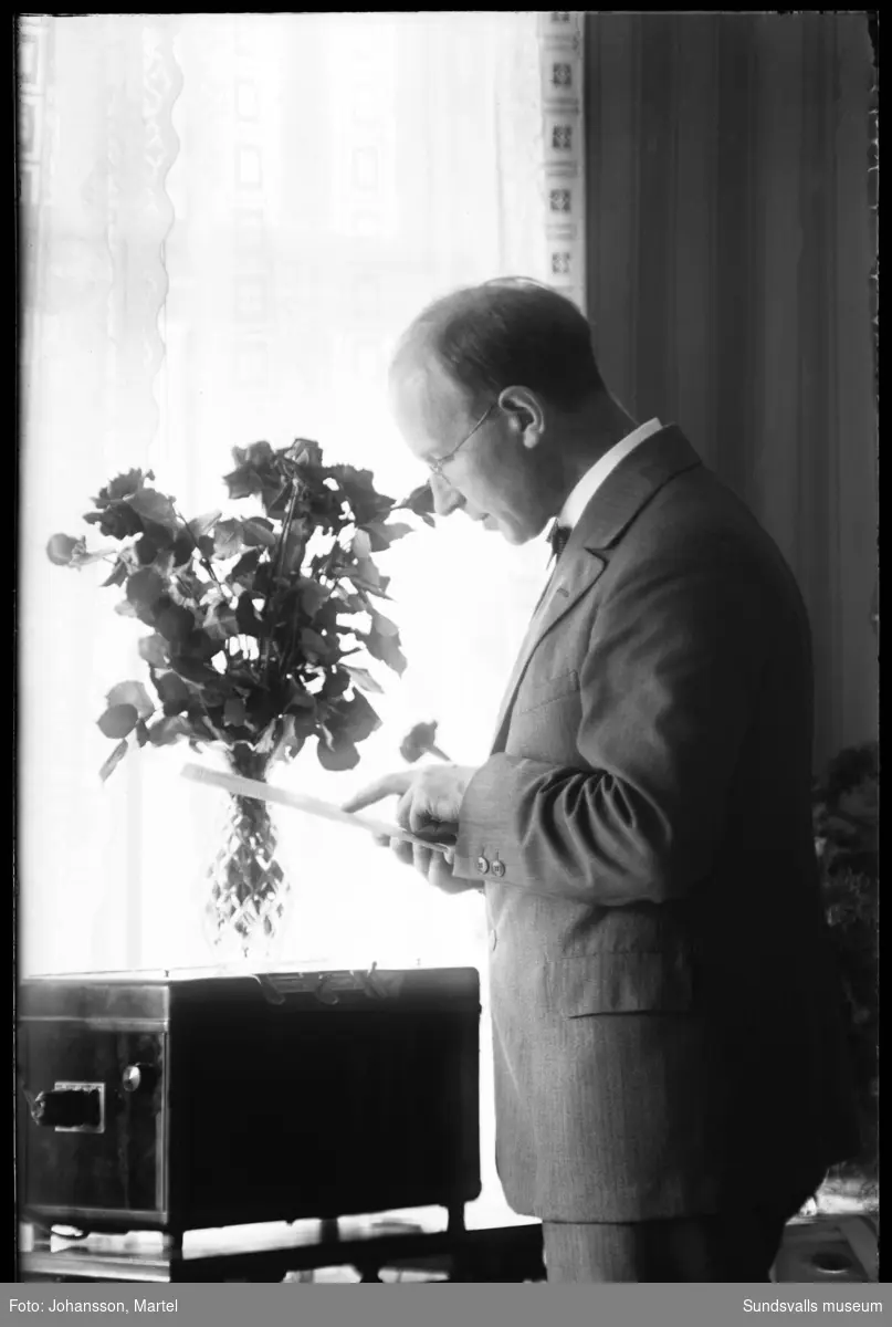 Martel Johansson står vid fönstret och läser på ett papper. På bordet står någon slags elektrisk apparat.