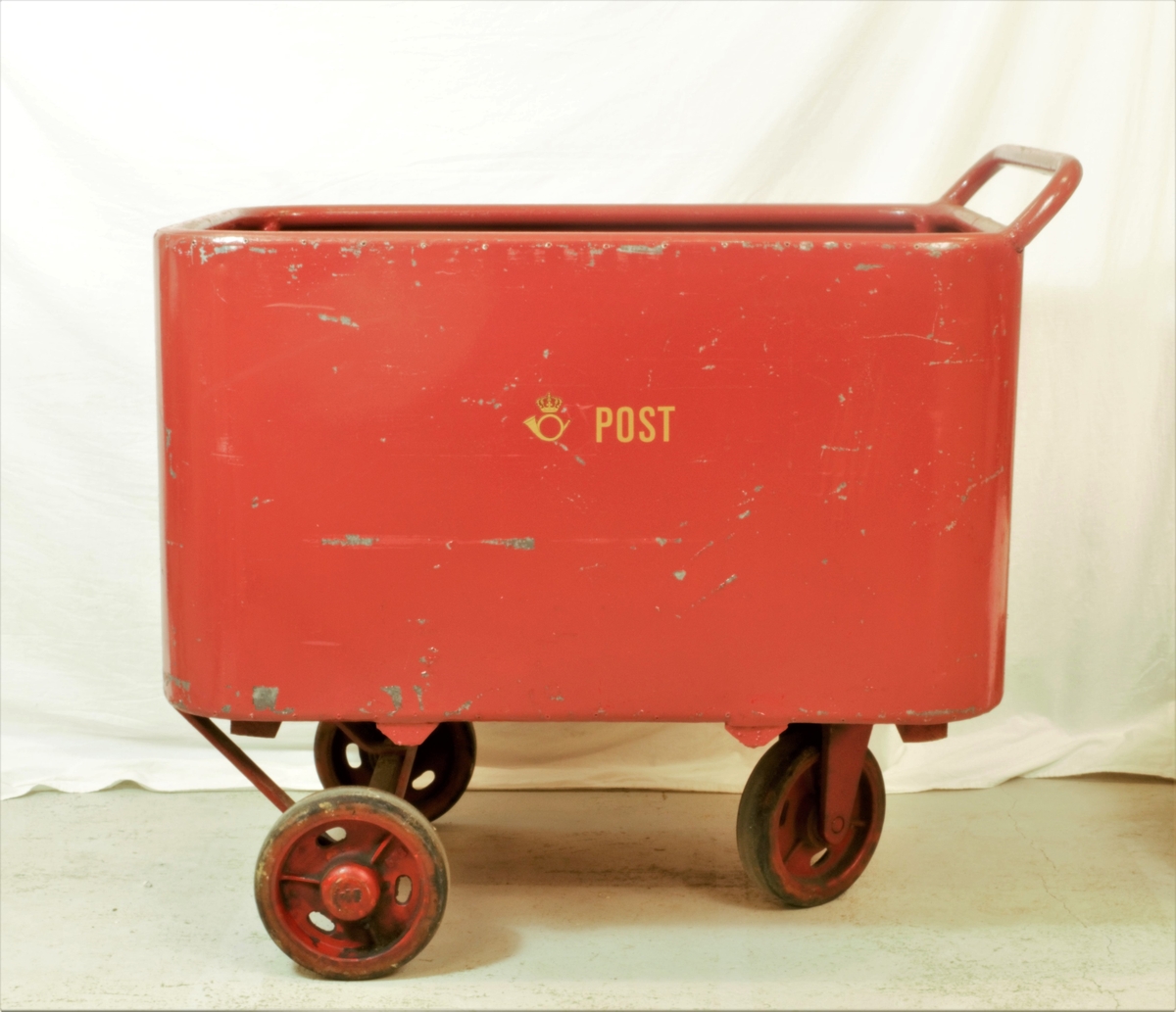 Rød postvogn med hvit logo. Selve vognen er i metall med tre gummibelagte hjul. Det er treplanker i bunnen av vognen.