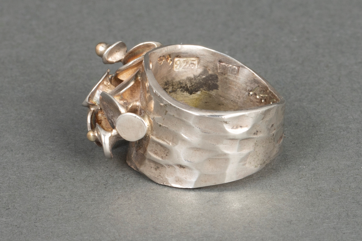 Bred, hamret fingerring med uregelmessig form og en rekke påloddede små, skråttstilte plater og tre små kuler av gull på oversiden.