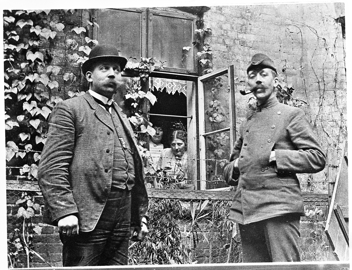 Gustav og Johan Skinderviken står foran en husvegg, mens Ragnhild Sveen Rudi og Oline Sveen ser av ut av vinduet bak. Bildet er tatt 9/4-1908.