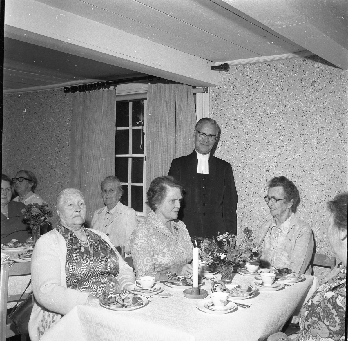 En präst står bland äldre kvinnor som sitter vid kaffebord, de samtalar och skrattar. Ett ljus är tänt på bordet.
