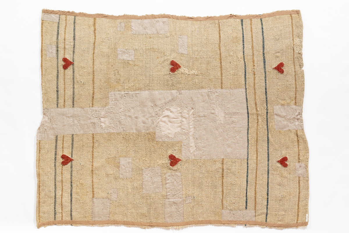 Filt (täcke) av naturfärgad ull vävd i tuskaft. Inslag i blågrått, gult och beige. Filten är dekorerad med "JPS 1745" och påsydda röda hjärtan av kläde.