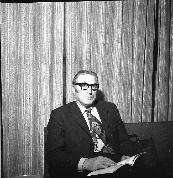 En man med kostym och mönstrad slips sitter med en bok i knät.