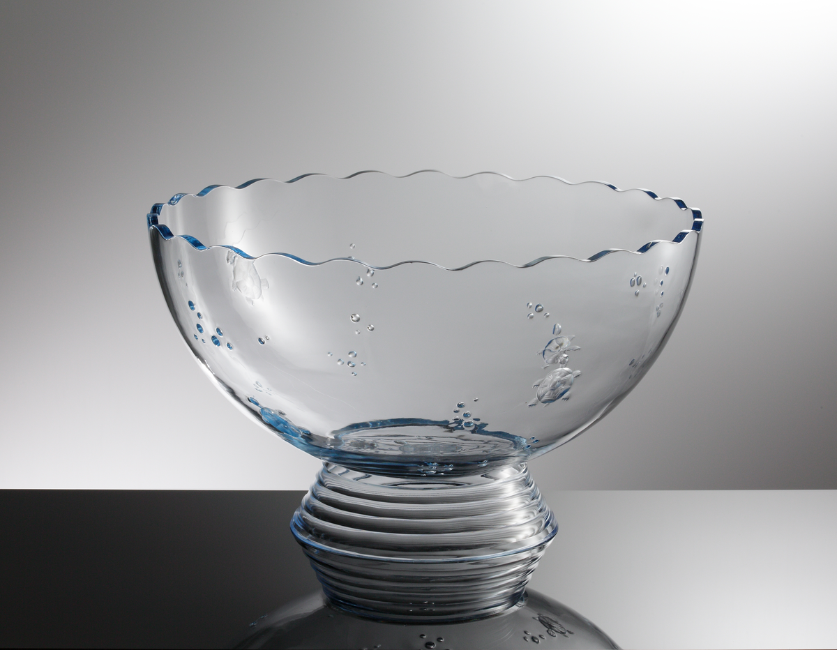 Formgiven av Edward Hald. Stor skål i ljusblå glasmassa med lös fotklack. Halvklotsformad med vågig mynningkant. Graverade sköldpaddor och luftbubblor som dekor. Konande fot med fasettslipade centriska ringar.