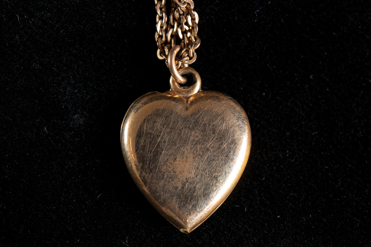Halskedja i guldfärgad metall med hjärtformad berlock. Berlockens framsida graverad och ornerad samt försedd med en infattad pärla.