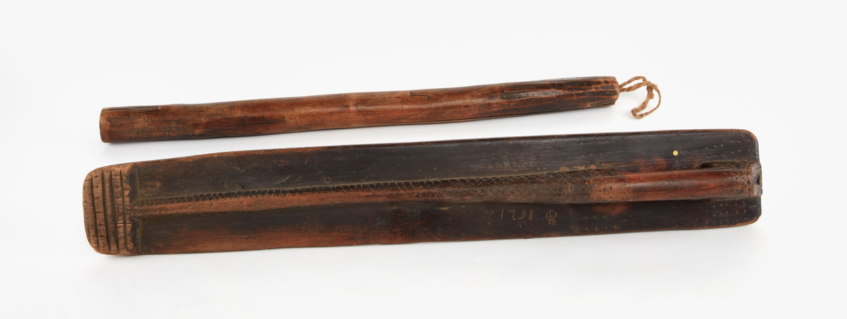 Mangeldon av trä med kavel. Handtag utsparat i ett stycke med brädan. Handtagets förlängning bildar mittås, som har ristad ornering i rutmönster. 
Märkt med initialer och bomärke samt år 1791. Rak typ.