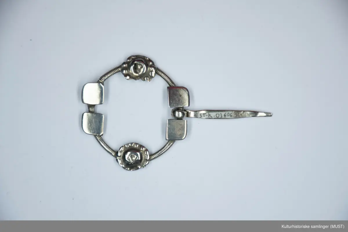 Ringen er av snodd sølv og  en nål er festet på tvers. På ringen er det fire plater og to blomsterornamenter. 