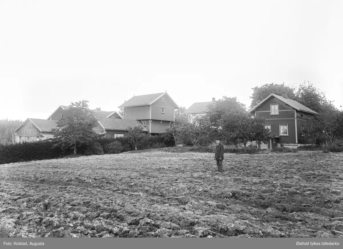 Bonde poserer i åkeren foran gårdsbruket, med våningshus, stabbur og uthus. Ukjent sted og person.