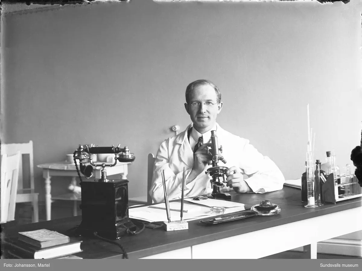 Doktor Martel Johansson sitter bakom skrivbordet med ett mikroskop på sin läkarmottagning. Okänt var i Sverige bilden är tagen.